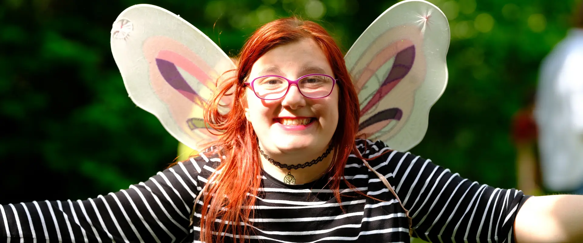 Mädchen lacht mit ausgebreiteten Armen in die Kamera, sie trägt Schmetterlingsflügel